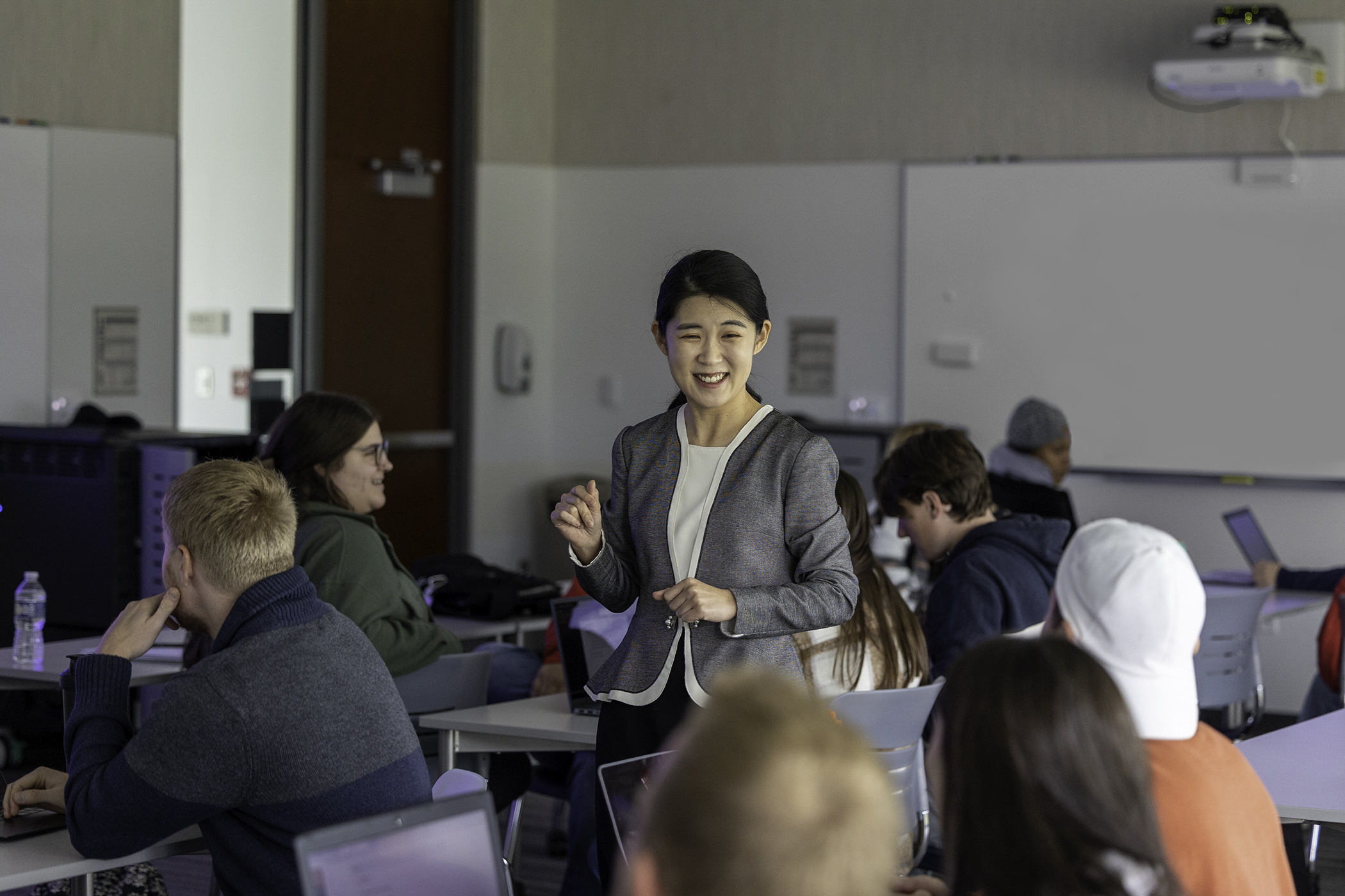 dark-haired woman in blazer stands among seated college students